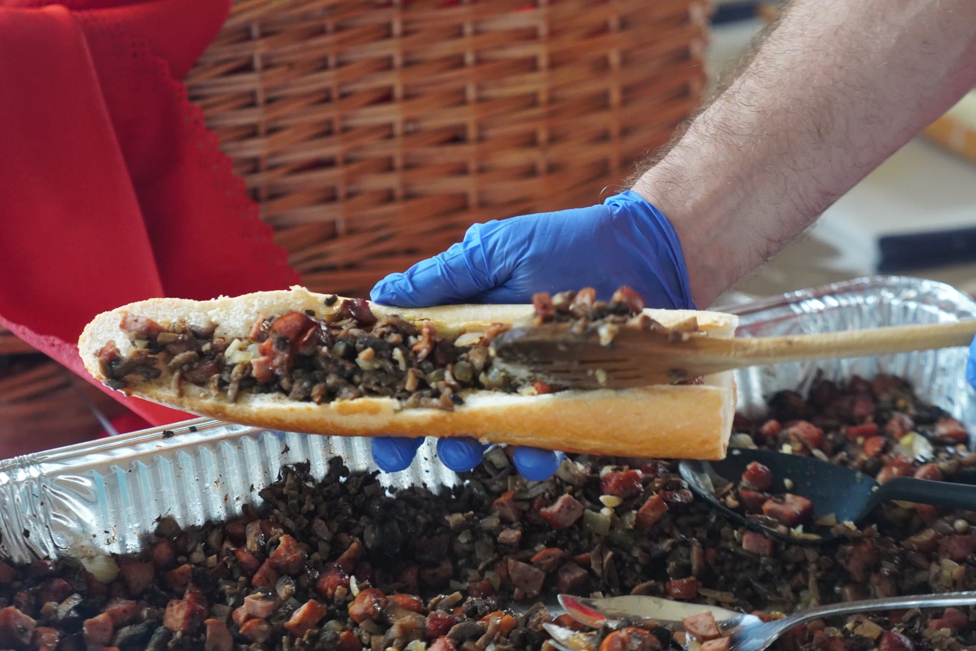 A gloved hand lifting a long zapiekanka roll, loaded with mushrooms, out of a foil tray heaped with more of the filling.