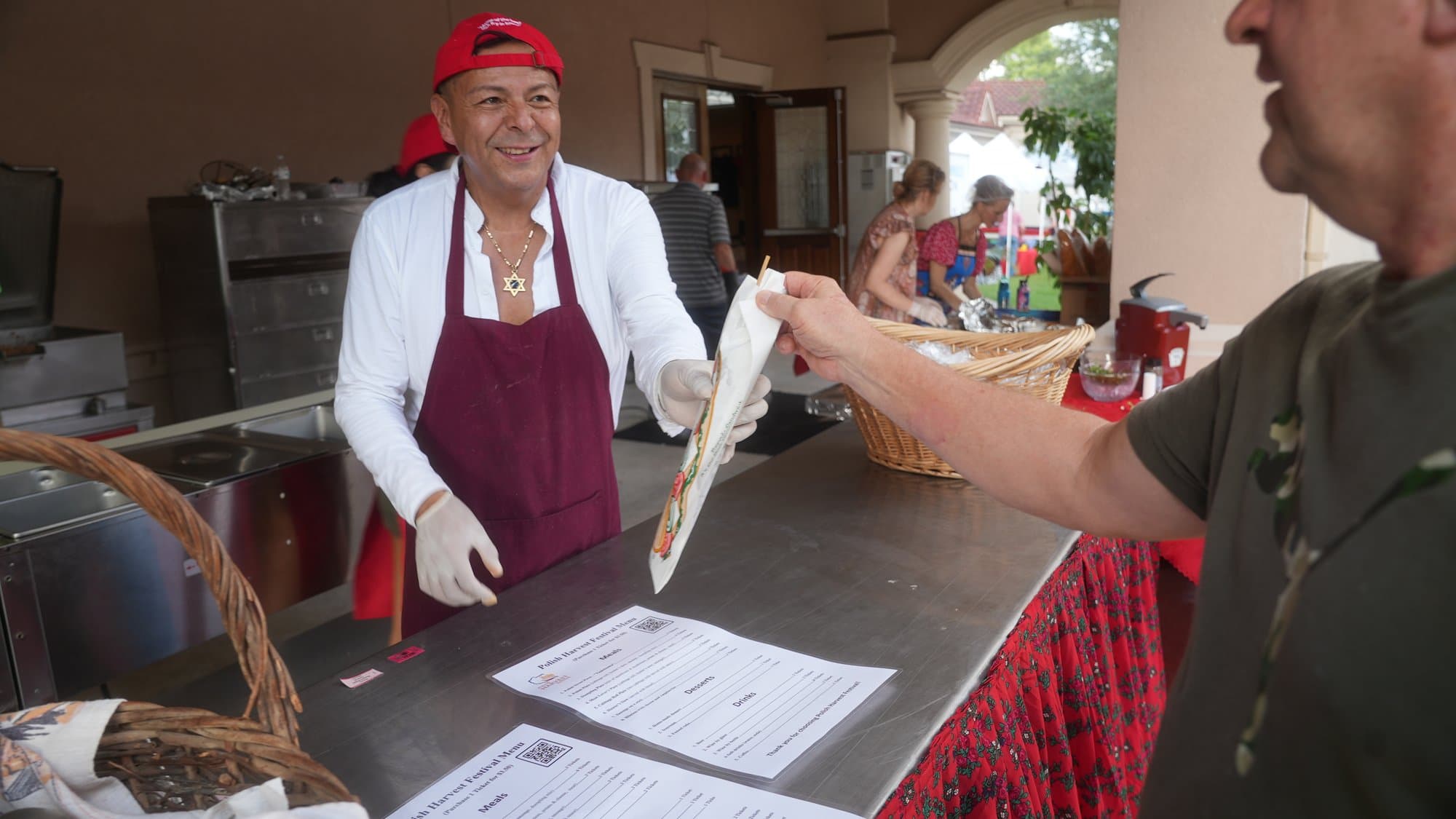 A volunteer in a red apron and white serving gloves piping cream into pastries at the counter, a wicker basket beside her and a customer's hand reaching in.