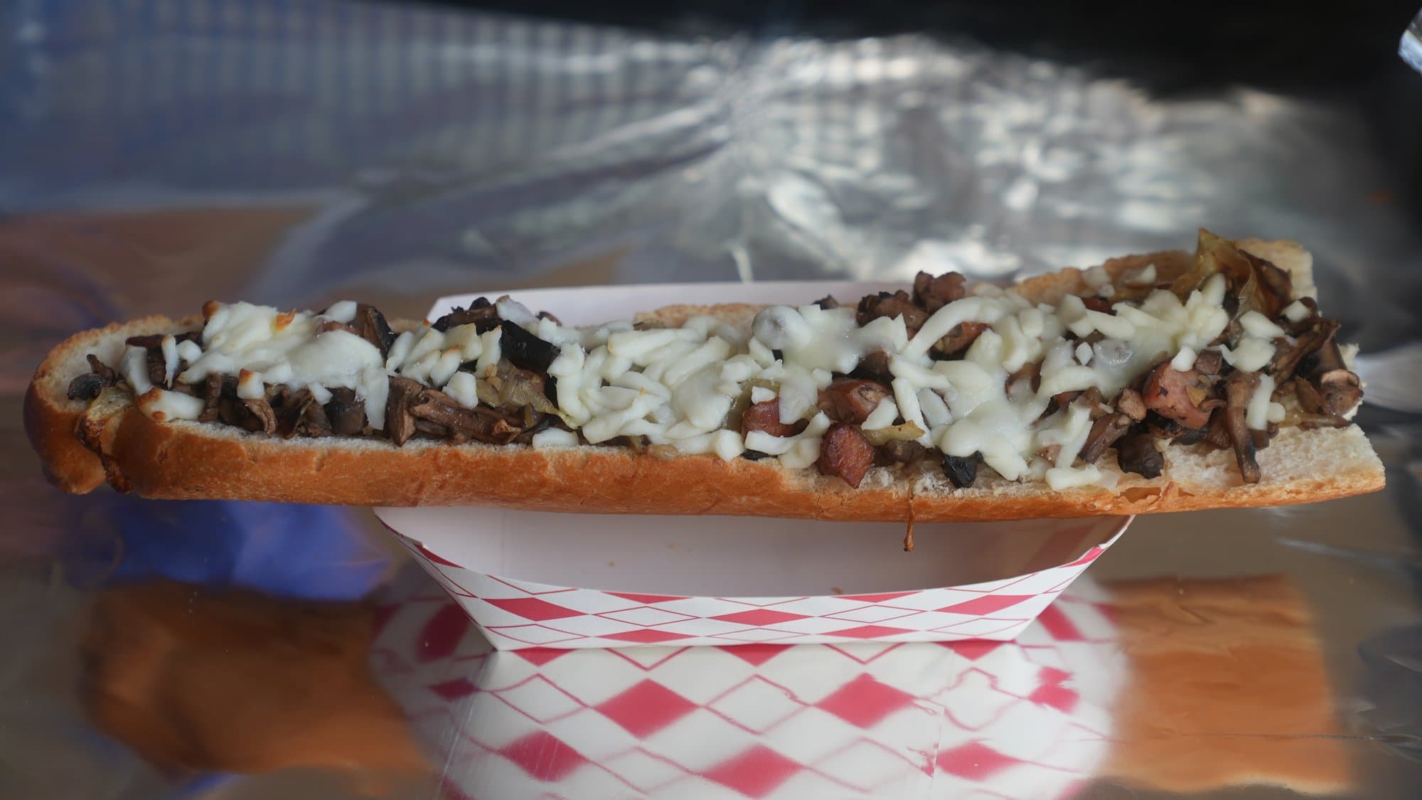 A zapiekanka close up: a long open-faced roll heaped with chopped mushrooms and melted white cheese, served in a red and white checked paper tray.